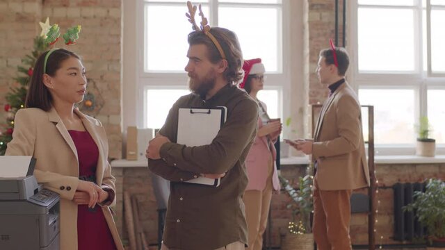 PAN portrait shot of cheerful bearded businessman and his female colleague in festive headbands standing by copier in office on Christmas and chatting, then looking at camera