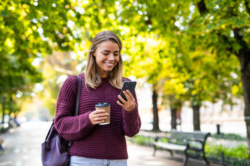 Beautiful young woman w carrying backpack walking outdoors, using mobile phone, holding coffee cup