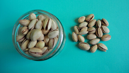 piled pistachio seeds next to a full glass jar of pistachios on a turquoise background top view