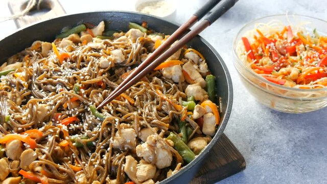 Japanese soba rice noodles with pieces of chicken, carrot, green shoots, sesame seeds, soy sauce. National food in a restaurant in large frying pan next to wooden bamboo sticks. Glass noodle salad.