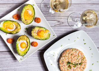 Aguacates con uevo al horno y tartar de salmón con dos copas de vino blanco en un fondo gris