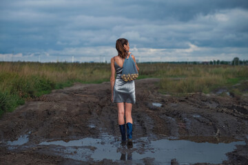 Fashion model woman holding string bag with potatoes, stay in dirty puddle in field, she is wearing a silver short dress and blue rubber boots. Concept of village style.