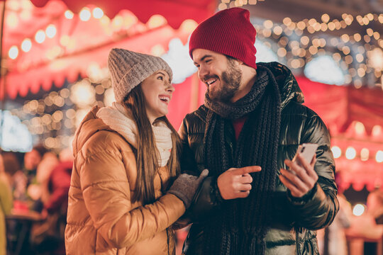 Photo of two people couple students have fun city christmas fair guy point index finger smartphone show post around evening x-mas illumination lights outside