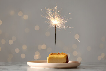 Piece of delicious cake with burning sparkler on white marble table against blurred lights