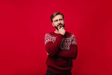Cheerful man with brown shiny hair standing on red background. Dreamy european guy in new year sweater chilling in studio.