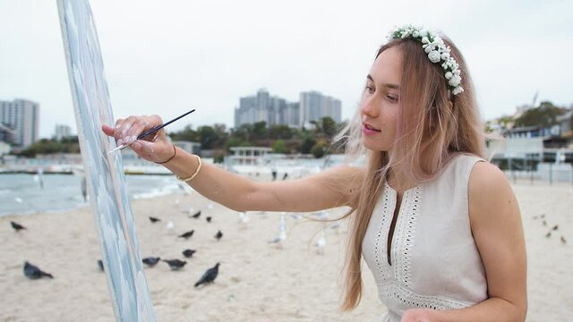 Young blond woman paints a picture on the beach. On open air. Against the background of flying seagulls. Slowmotion