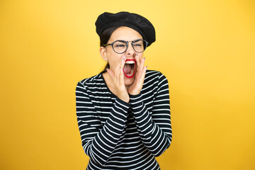 Young beautiful brunette woman wearing french beret and glasses over yellow background shouting and screaming loud to side with hands on mouth