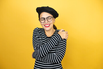 Young beautiful brunette woman wearing french beret and glasses over yellow background hugging...