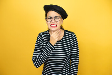 Young beautiful brunette woman wearing french beret and glasses over yellow background touching painful neck, sore throat for flu, clod and infection