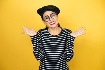 Young beautiful brunette woman wearing french beret and glasses over yellow background clueless and confused expression with arms and hands raised