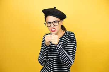 Young beautiful brunette woman wearing french beret and glasses over yellow background Punching fist to fight, aggressive and angry attack, threat and violence