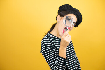 Young beautiful brunette woman wearing french beret and glasses over yellow background surprised...