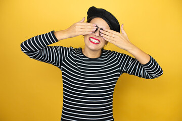 Young beautiful brunette woman wearing french beret and glasses over yellow background covering eyes with hands smiling cheerful and funny. Blind concept.