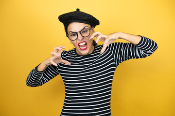 Young beautiful brunette woman wearing french beret and glasses over yellow background crazy and mad shouting and yelling with aggressive expression and arms raised. Frustration concept.