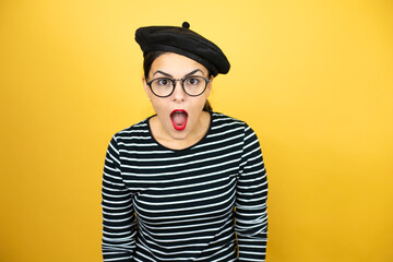 Young beautiful brunette woman wearing french beret and glasses over yellow background afraid and shocked with surprise and amazed expression, fear and excited face.