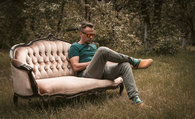 Obraz premium Resting man in grey jeans sitting on a sofa in the middle of garden. High quality photo