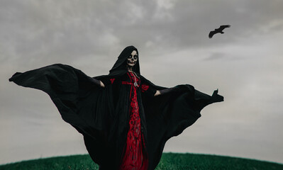 Woman stands among field in Halloween costume of death against dark sky and flying raven.