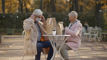 Portrait of senior couple putting on safety mask leaving outdoors cafe with takeaway order - Powered by Adobe