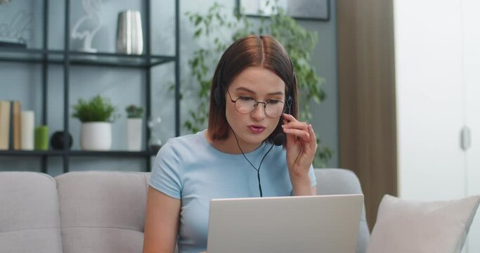 Concentrated customer support representative in headphones working while sitting on sofa at home. Good-looking young woman in glasses talking to client via video call. Service concept.