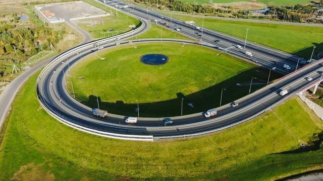 Cars drive along modern highway with round road junction and overpass near green meadow at bright sunlight aerial view timelapse