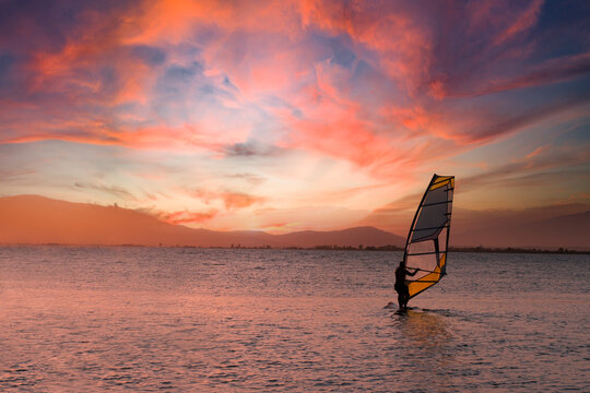 Windsurfer riding on a surfboard against a cloudy orange sunset. Empty copy space for Editor's content.