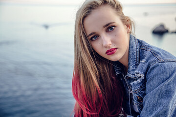 Close portrait young girl in front of the sea in stylish clothes with hairstyle and makeup. Background blurred for artistic purposes.