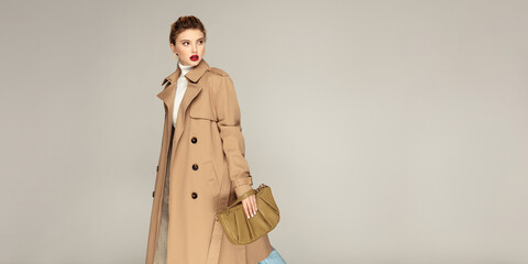 Autumn clothing concept. Fashionable young model posing against grey background dressed in trench coat and holding stylish  bag.
