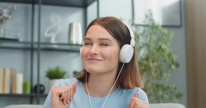 Portrait of charming Caucasian woman having fun while listening to music at home. Attractive young female in headphones dacing and singing in living room. Entertainment, leisure time concept.