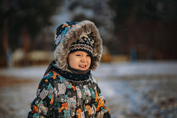 little cute caucasian boy walking on the snowy beach squinting into the sun. Kid is wearing winter warm jacket with hood on, knit hat and a scarf