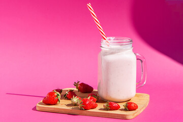 Strawberry smoothie in a glass jar with a red-striped tube. There are strawberries on a wooden Board and a strawberry smoothie next to them. Concept of healthy food. On a bright pink background