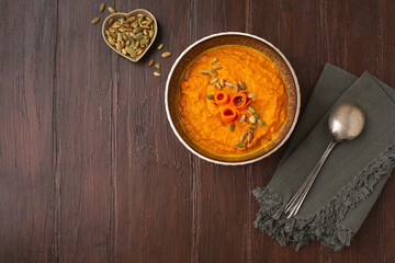 Top View of Homemade Carrot Squash in a Bowl on a Wooden Table; Garnished with Carrots and Pumpkin Seeds; Spoon and Napkin Beside Bowl
