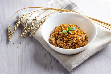Spelt porridge in a bowl with basil leaves on linen napkin with ears of spelt grain. White wooden...