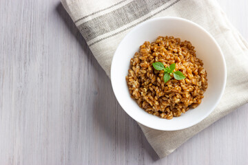 Spelt porridge in a bowl with basil on the linen napkin. White wooden background. Copy space, top view.
