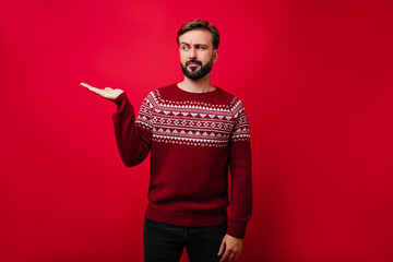 Pensive man in knitted christmas sweater posing in studio. Bearded guy standing on red background with hand up.
