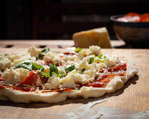 Raw pizza with salami, mozzarella and basil waiting for baking. Parmeggiano  cheese and tomatoes in a bowl in the background