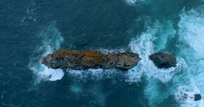 Aerial vertical view of a separate, tall, narrow reef in the ocean, surrounded by surfing waves. Slow Motion, 4K drone footage of Madeira atlantic coast. Madeira nature, Portugal.