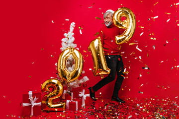 Emotional old man dancing under confetti at new year party. Studio portrait of enthusiastic santa claus posing on red background.