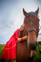 Obraz premium A woman in a bright Gypsy dress and image with a horse in a field with green grass. A model or actress posing in nature with an animal from a farm and the sky with clouds in the background