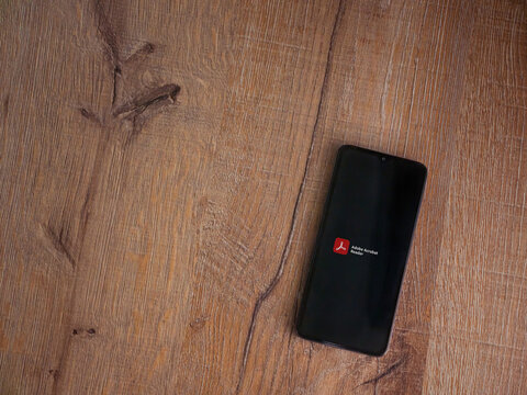 Lod, Israel - July 8, 2020: Adobe Acrobat Reader app launch screen with logo on the display of a black mobile smartphone on wooden background. Top view flat lay with copy space.