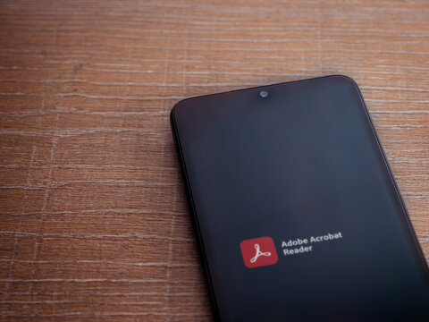 Lod, Israel - July 8, 2020: Adobe Acrobat Reader app launch screen with logo on the display of a black mobile smartphone on wooden background. Top view flat lay with copy space.