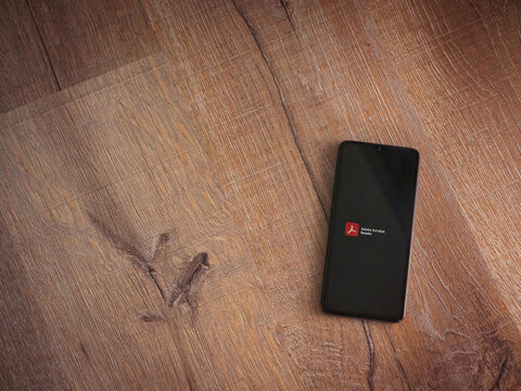 Lod, Israel - July 8, 2020: Adobe Acrobat Reader app launch screen with logo on the display of a black mobile smartphone on wooden background. Top view flat lay with copy space.