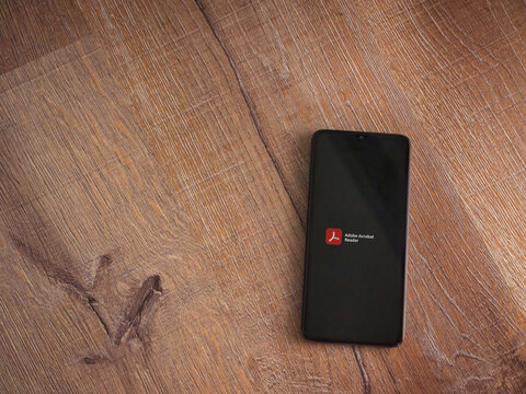 Lod, Israel - July 8, 2020: Adobe Acrobat Reader app launch screen with logo on the display of a black mobile smartphone on wooden background. Top view flat lay with copy space.