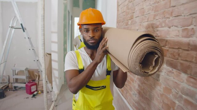 Portrait of african builder in safety helmet carrying roll of construction material indoors