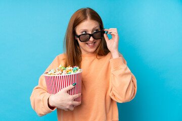 Obraz premium Young redhead woman over isolated background with 3d glasses and holding a big bucket of popcorns