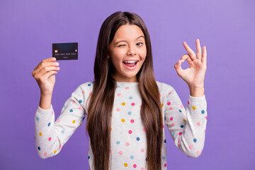 Photo portrait of cheerful small girl demonstrating bank card showing okay sign winking blinking...