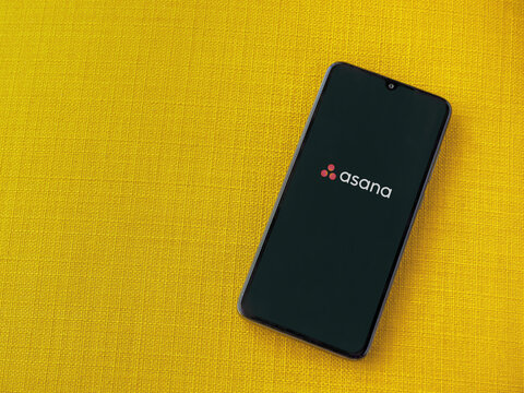 Lod, Israel - July 8, 2020: Asana app launch screen with logo on the display of a black mobile smartphone on a yellow fabric background. Top view flat lay with copy space.