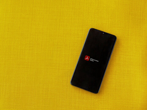 Lod, Israel - July 8, 2020: Adobe Acrobat Reader app launch screen with logo on the display of a black mobile smartphone on a yellow fabric background. Top view flat lay with copy space.