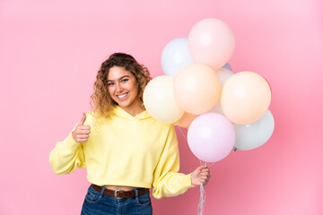 Young blonde woman with curly hair catching many balloons isolated on pink background giving a thumbs up gesture