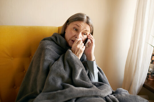 Tired sick senior woman having cold, blowing nose in tissue, sitting on couch with mobile, calling doctor or relative, suffering from headache, fever or high temperature. Technology, age and health - Powered by Adobe