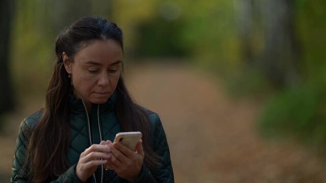 middle-aged woman is walking in park at autumn day, reading messages in smartphone and smiling
